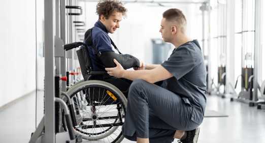 chłopak na wózku z ręką w temblaku, fizjoterapeuta bada jego kończynę